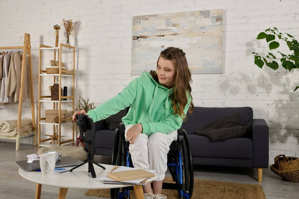 Uzun kahverengi saçlı genç bir kadın oturma odasında tekerlekli sandalyede oturuyor, önünde bir telefon tutuyor ve video çekiyor.. - Fotoğraf, Görsel