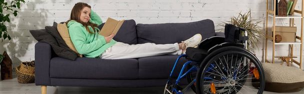 Brunetka młoda kobieta siedzi na wózku inwalidzkim, spoczywa na kanapie czytając książkę w salonie. - Zdjęcie, obraz