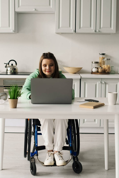 Egy fiatal, fogyatékos barna nő tolószékben ül a konyhájában, és laptopot használ.. - Fotó, kép