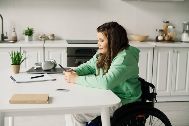 Une femme brune est assise dans un fauteuil roulant à la maison, utilisant son téléphone tout en travaillant à une table. - Photo, image