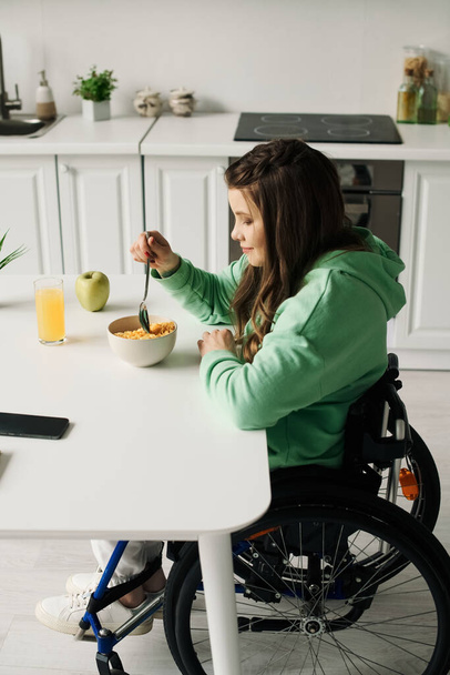 Młoda brunetka na wózku inwalidzkim siedzi przy stole w salonie, delektując się miską płatków śniadaniowych i szklanką soku pomarańczowego. - Zdjęcie, obraz