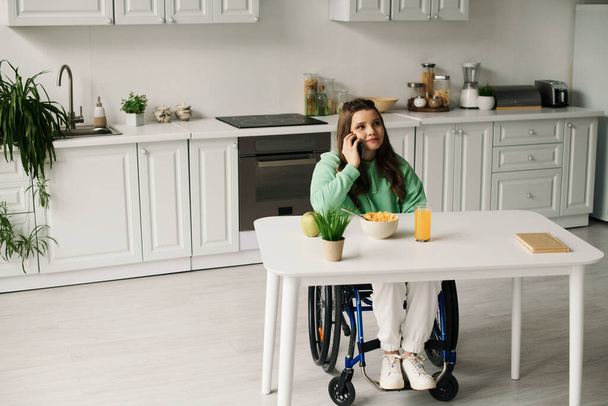 Μια νεαρή μελαχρινή γυναίκα σε αναπηρική καρέκλα κάθεται σε ένα τραπέζι, μιλώντας στο τηλέφωνό της. - Φωτογραφία, εικόνα
