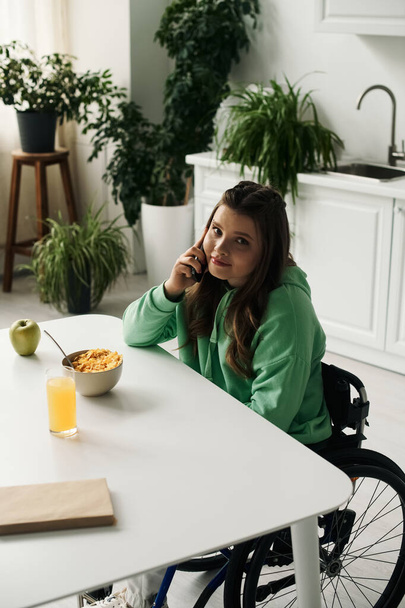 Uma jovem morena com deficiência senta-se em uma cadeira de rodas em sua mesa de cozinha, conversando em seu telefone celular. - Foto, Imagem