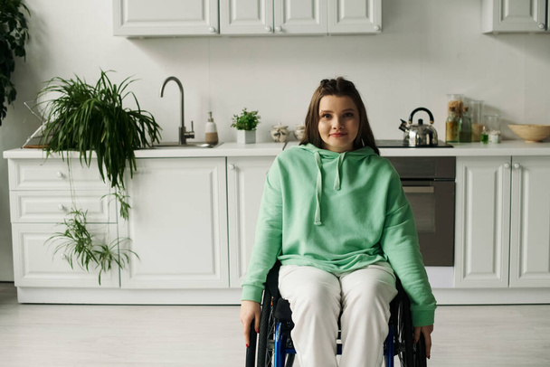 Brunetka młoda kobieta na wózku inwalidzkim siedzi w kuchni i uśmiecha się do kamery. - Zdjęcie, obraz