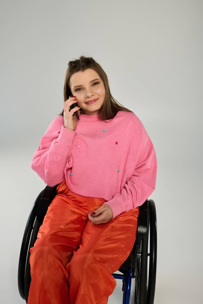 Mladá brunetka v růžovém svetru a oranžových kalhotách sedí na vozíčku a mluví po telefonu. - Fotografie, Obrázek