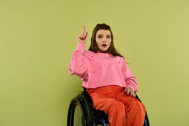 Młoda kobieta na wózku inwalidzkim siedzi w studio, wskazując w górę z zaskakującym wyrazem twarzy.. - Zdjęcie, obraz