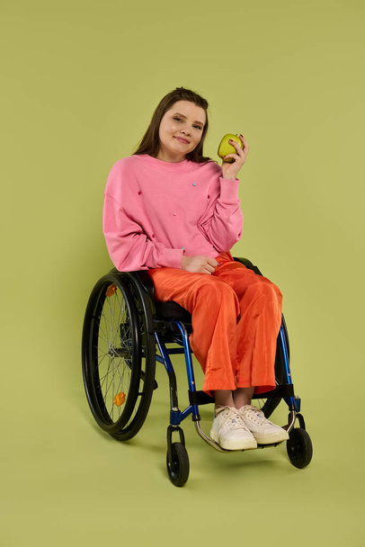 Une jeune femme en fauteuil roulant est assise dans un studio, tenant une pomme et regardant directement la caméra. Elle porte des vêtements décontractés et un sourire. - Photo, image