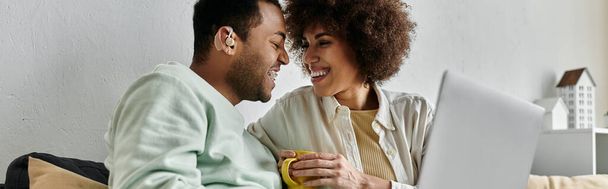 Femme afro-américaine joyeuse assise près de son mari avec une aide auditive et regardant un ordinateur portable. - Photo, image