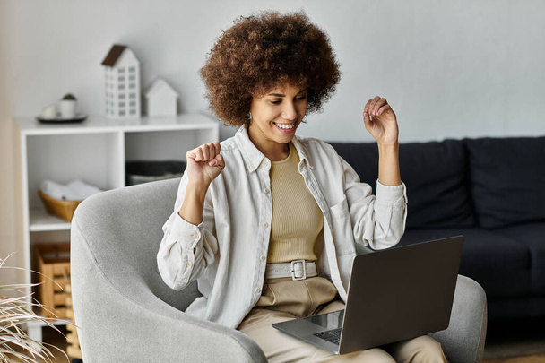 Una joven y atractiva mujer afroamericana se sienta en una silla, sonriendo amplia y excitadamente usando lenguaje de señas, junto a la computadora portátil. - Foto, imagen