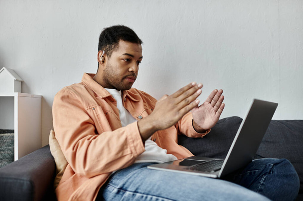 Un jeune Afro-Américain assis sur un canapé, utilisant la langue des signes tout en regardant un ordinateur portable. - Photo, image