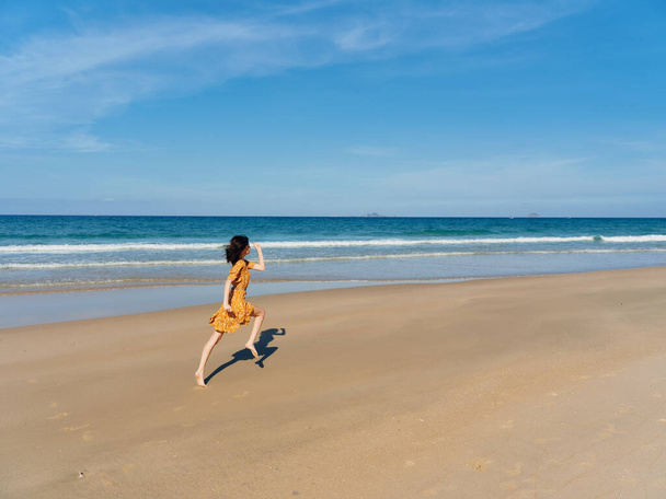 Mujer asiática alegre en un fluido vestido amarillo corriendo enérgicamente en la playa de arena bajo un cielo azul claro - Foto, imagen