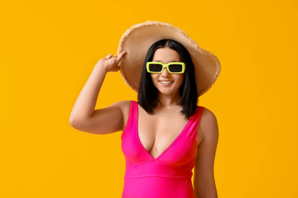 Beautiful young woman in stylish pink swimsuit and sunglasses on yellow background - Photo, Image