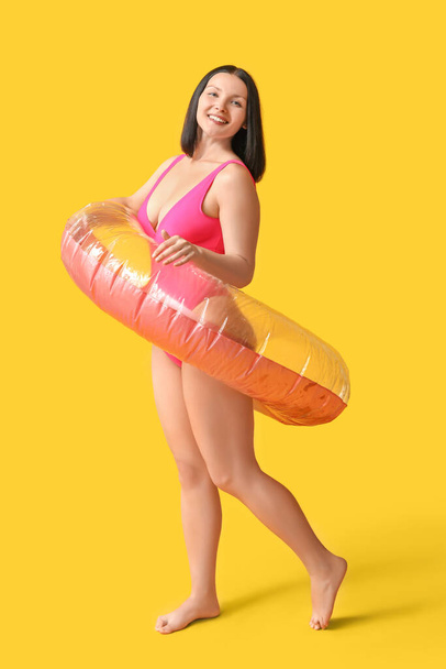 Beautiful young woman in stylish pink swimsuit with inflatable ring on yellow background - Photo, Image