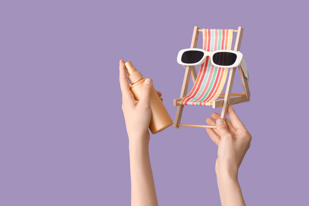 Mains féminines avec crème solaire et mini chaise longue avec lunettes de soleil sur fond lilas - Photo, image