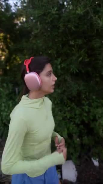 Una mujer en ropa deportiva y auriculares rosados está trotando a través de un parque, disfrutando de la vegetación y los árboles a su alrededor - Metraje, vídeo