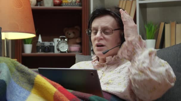Starsza kobieta, leżąca na kanapie, w domu, przykryta ciepłym kocem, komunikuje się z wnukiem, elektronicznie, za pomocą tabletu, uśmiechając się słodko podczas rozmowy. - Materiał filmowy, wideo