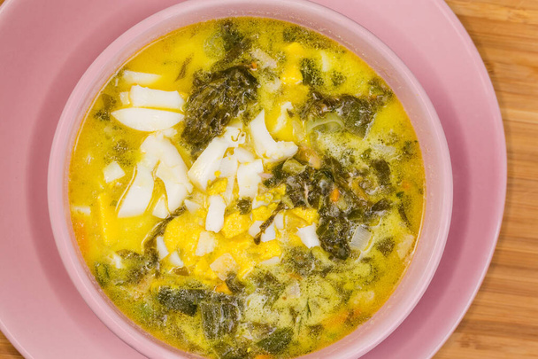 Σερβίρισμα της σούπας σορέλ, επίσης γνωστή ως πράσινο μπορς με ψιλοκομμένο βραστό αυγό και ξινή κρέμα σε ένα ροζ μπολ σε ένα ξύλινο τραπέζι, top view close-up - Φωτογραφία, εικόνα