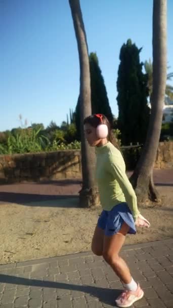 Жінка біжить у сонячному парку, одягнена в зелений верх, сині шорти та рожеве взуття, одягнені в навушники - Кадри, відео