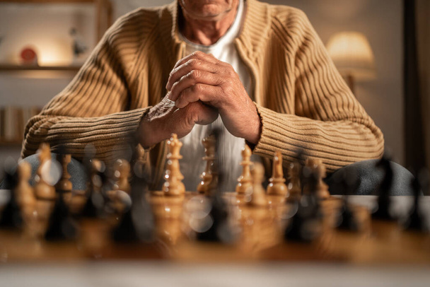 Serieuze senior man zit in de avondwoonkamer en schaken in eenzaamheid. Eenzaamheid en pensioen concept. Kopieerruimte - Foto, afbeelding