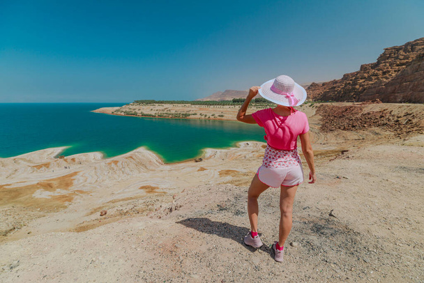 Elegant woman in summer dress holding a hat, gazing at the serene waters of the Dead Sea under a bright blue sky in Jordan - Photo, Image