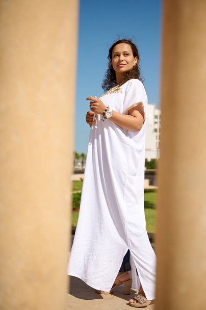 Selbstbewusste Frau im weißen Kleid, die an einem sonnigen Tag im Freien spazieren geht. Sie wirkt entspannt und stilvoll, verströmt ein Gefühl von Anmut und Eleganz vor dem Hintergrund von Säulen und einem klaren blauen Himmel. - Foto, Bild