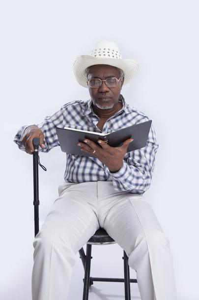 Hombre de edad en sombrero con bastón se sienta en la silla y lee libro
 - Foto, Imagen