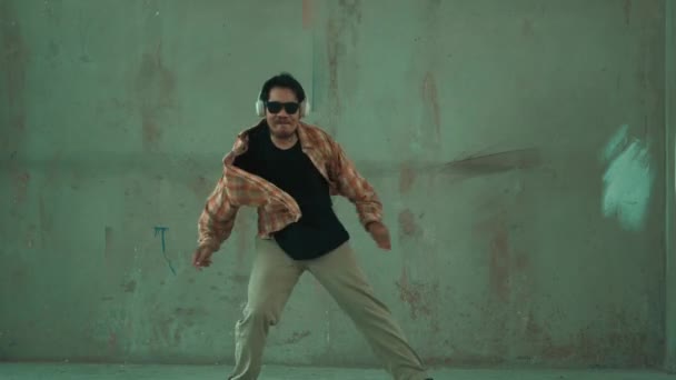 Aantrekkelijke Latijns-Amerikaanse danser in casual look praktijk straat dans, terwijl muziek luisteren vanaf de hoofdtelefoon op cement achtergrond. Motion shot van breakdancevoorstelling. Outdoor sport 2024. hiphop. - Video