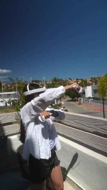 Žena zažívá virtuální realitu za slunečného dne z balkónu, pomocí soupravy VR a ovladačů. - Záběry, video