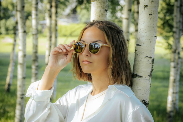 Une femme élégante dans une chemise blanche ajuste ses lunettes de soleil tout en se tenant dans une forêt de bouleaux. La lumière du soleil et le fond naturel réfléchissent ses lunettes, créant un look cool et à la mode. - Photo, image