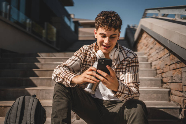 Giovane ragazzo con cuffie e thermos tazza sedersi sulle scale e utilizzare il telefono cellulare - Foto, immagini