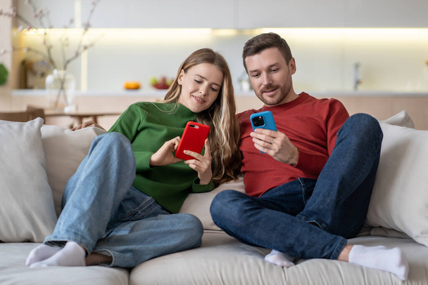 Joyeux couple heureux assis sur un canapé avec des smartphones, souriant, jouant à un jeu en ligne absorbant, regardant de courtes vidéos drôles sur Internet, échangeant des photos, ayant un appel vidéo de groupe avec des amis - Photo, image