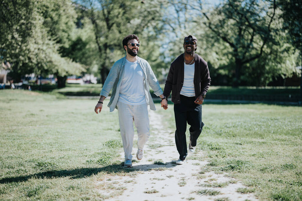 Ένα νεαρό, πολυφυλετικό γκέι ζευγάρι που κρατιέται χέρι-χέρι, περπατώντας χαρούμενα μέσα από ένα ηλιόλουστο πάρκο, δείχνοντας αγάπη και ευτυχία.. - Φωτογραφία, εικόνα