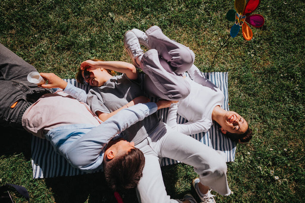 Три друга наслаждаются расслабляющим днем вместе, лежа на одеяле для пикника в траве, под ярким солнцем, разделяя радость и комфорт. - Фото, изображение