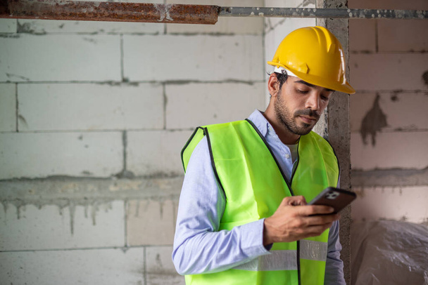 Człowiek pracownik budowlany nosić kamizelkę z żółtym hełmem za pomocą smartfona w czasie relaksu na placu budowy. mężczyzna pracownik wpisując dotyk czat na telefon komórkowy używać social media urządzenia komunikacja. - Zdjęcie, obraz
