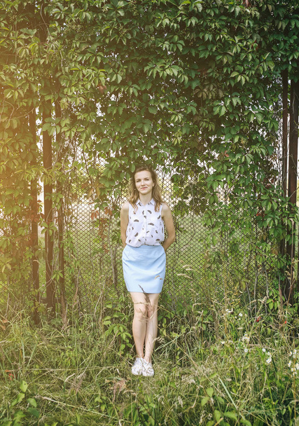 Дівчина-гіпстер стоїть біля паркану з листя плюща. Жінка в блакитній спідниці і блузці посміхається і насолоджується сонцем. Смішні і щасливі емоції в літній фотосесії. - Фото, зображення