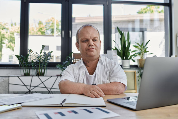 Un hombre con inclusividad se sienta en un escritorio en una oficina moderna, centrado en su trabajo. - Foto, imagen