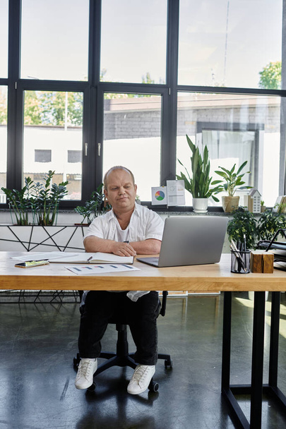 Un homme inclusif est assis à un bureau dans un environnement de bureau, travaillant sur un ordinateur portable. - Photo, image