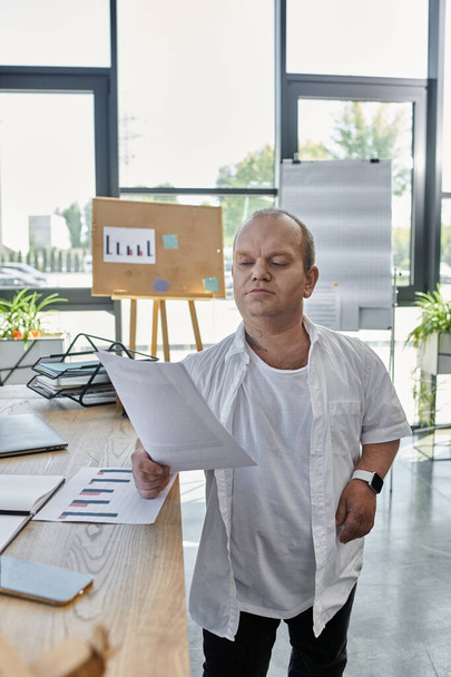 Ένας άντρας με εγκλειστικότητα σε λευκό πουκάμισο στέκεται δίπλα σε ένα γραφείο, εξετάζοντας έγγραφα.. - Φωτογραφία, εικόνα