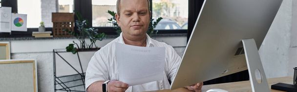 Ένας άντρας με εγκλειστικότητα κάθεται στο γραφείο του, εξετάζοντας έγγραφα σε ένα περιβάλλον γραφείου. - Φωτογραφία, εικόνα