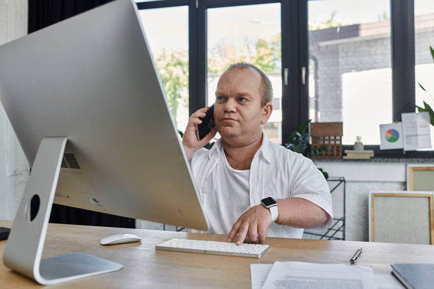 近代的なオフィスにいる男は,携帯電話で話している間,自分のコンピュータで働きます.. - 写真・画像