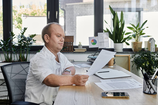 Een man met inclusiviteit zit aan zijn bureau in een goed verlicht kantoor en bekijkt documenten. - Foto, afbeelding