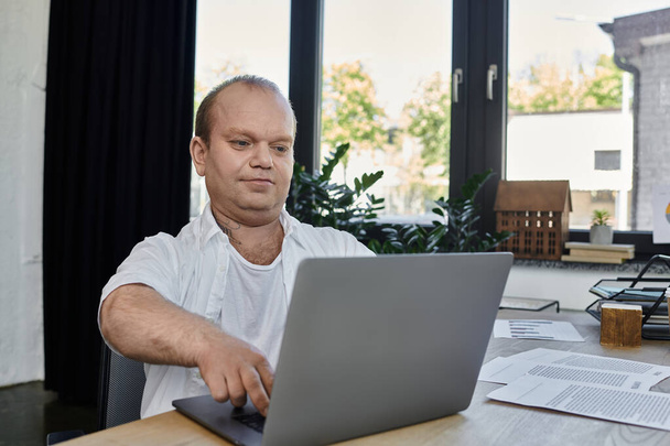 Ένας άντρας με εγκλειστικότητα κάθεται σε ένα γραφείο στο γραφείο του, δουλεύοντας εντατικά στο λάπτοπ του.. - Φωτογραφία, εικόνα