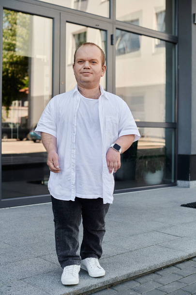 A man with inclusivity in a white shirt walks on a sidewalk outside a building with large glass doors. - Foto, imagen