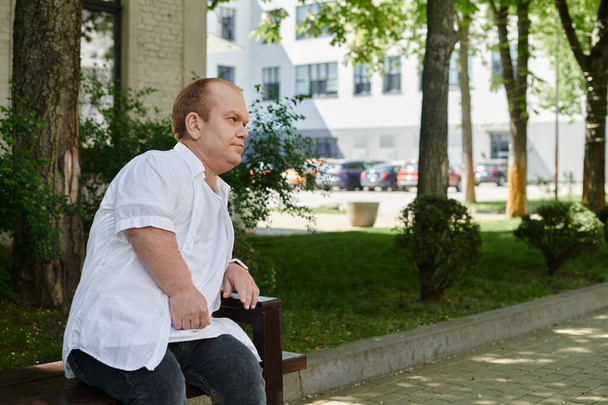 Ένας άντρας με εγκλειστικότητα κάθεται σε ένα παγκάκι σε ένα πάρκο της πόλης, περικυκλωμένος από πράσινο.. - Φωτογραφία, εικόνα