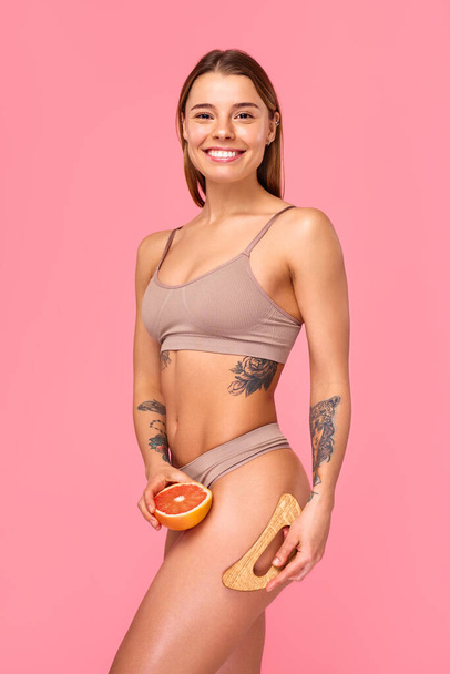 Женщину видят на снимке с грейпфрутом и отшелушивающей кисточкой, которая фокусируется на уходе за кожей, здоровье, самочувствии и красоте для продвижения здоровой и сияющей кожи на розовом фоне. - Фото, изображение