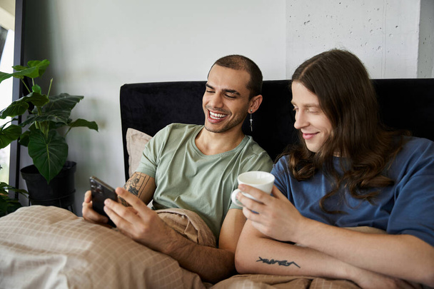 Een jong gay paar glimlacht en lacht samen in bed, genieten van hun ochtend koffie en kijken naar hun telefoon. - Foto, afbeelding