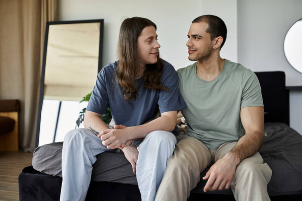 Mladý homosexuální pár sdílí něžný okamžik na posteli, oči upřené do láskyplného pohledu. - Fotografie, Obrázek
