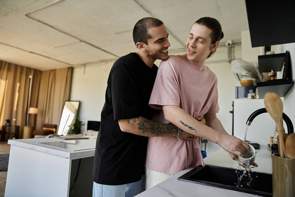 Een jong gay paar geniet van een moment van speelse intimiteit tijdens het wassen van de afwas in hun moderne keuken. - Foto, afbeelding