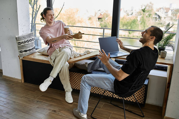 Un jeune couple gay profite d'une pause café dans leur maison moderne, riant et parlant pendant qu'on utilise un ordinateur portable. - Photo, image