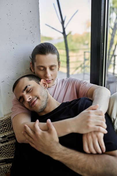 Twee jonge mannen knuffelen samen, ogen dicht, in een modern huis, genietend van een vredig moment van intimiteit. - Foto, afbeelding